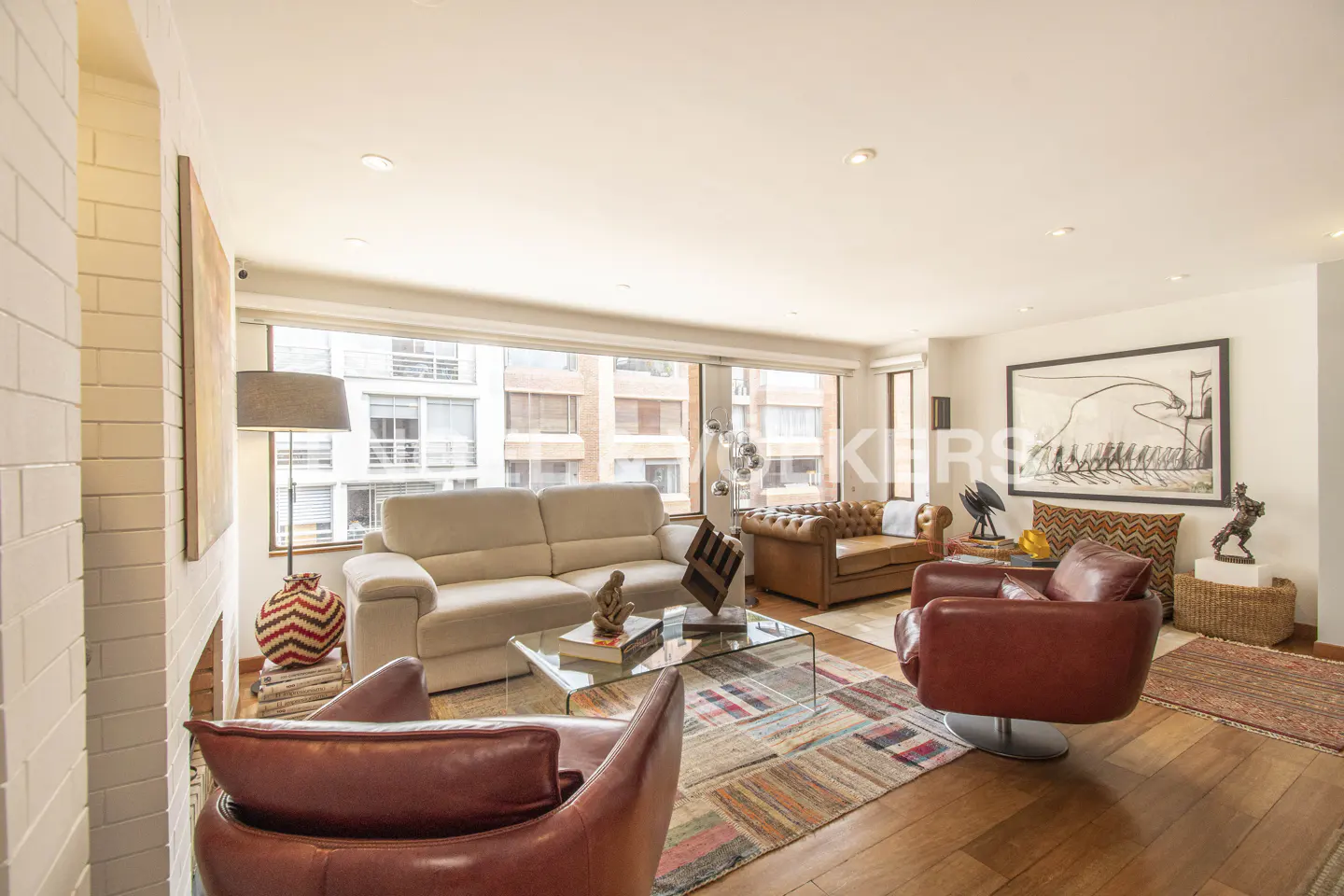 Bright living room with a cream sofa, two red leather armchairs, a glass coffee table, and a colorful patterned rug.