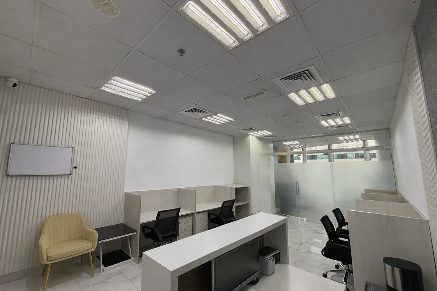 Modern office space with white walls, cubicle desks, black chairs, a beige armchair, and a whiteboard on a ribbed wall.