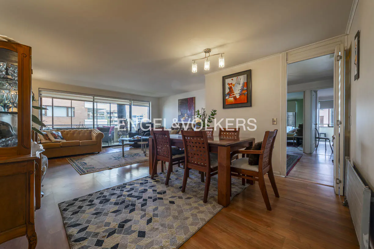 A bright living and dining area with a wooden table, chairs, and a patterned rug. A tan sofa is visible through a large window.