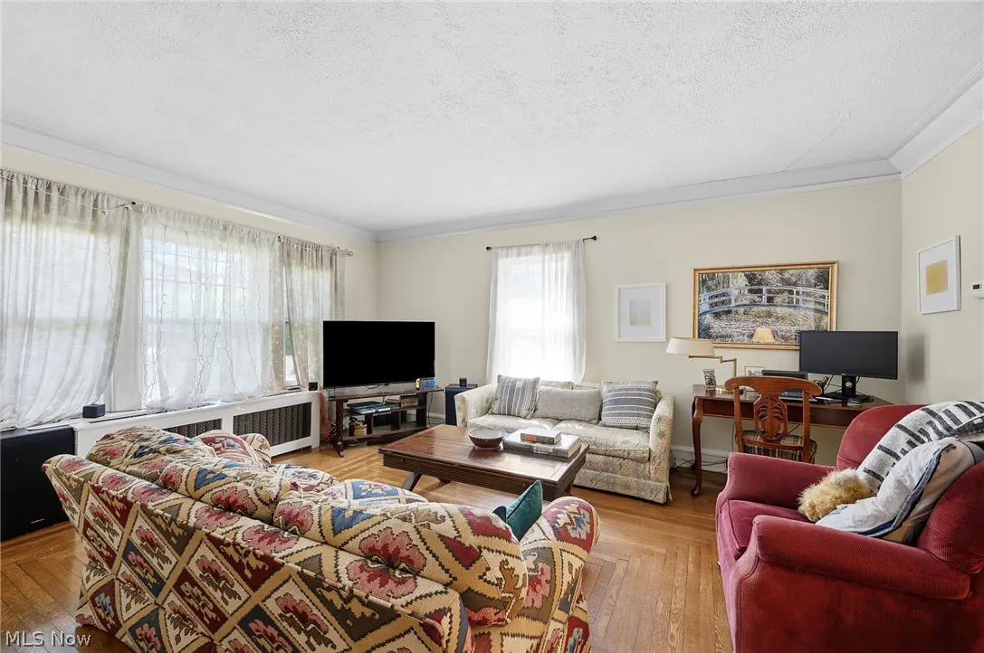 Living room with patterned sofas, a red armchair, and a wooden desk with a computer. Natural light filters through sheer curtains.