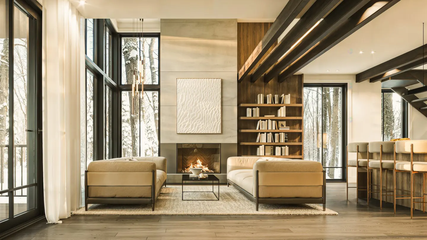 Bright living room with beige sofas facing a fireplace, large windows showing a snowy forest, and a bookshelf.
