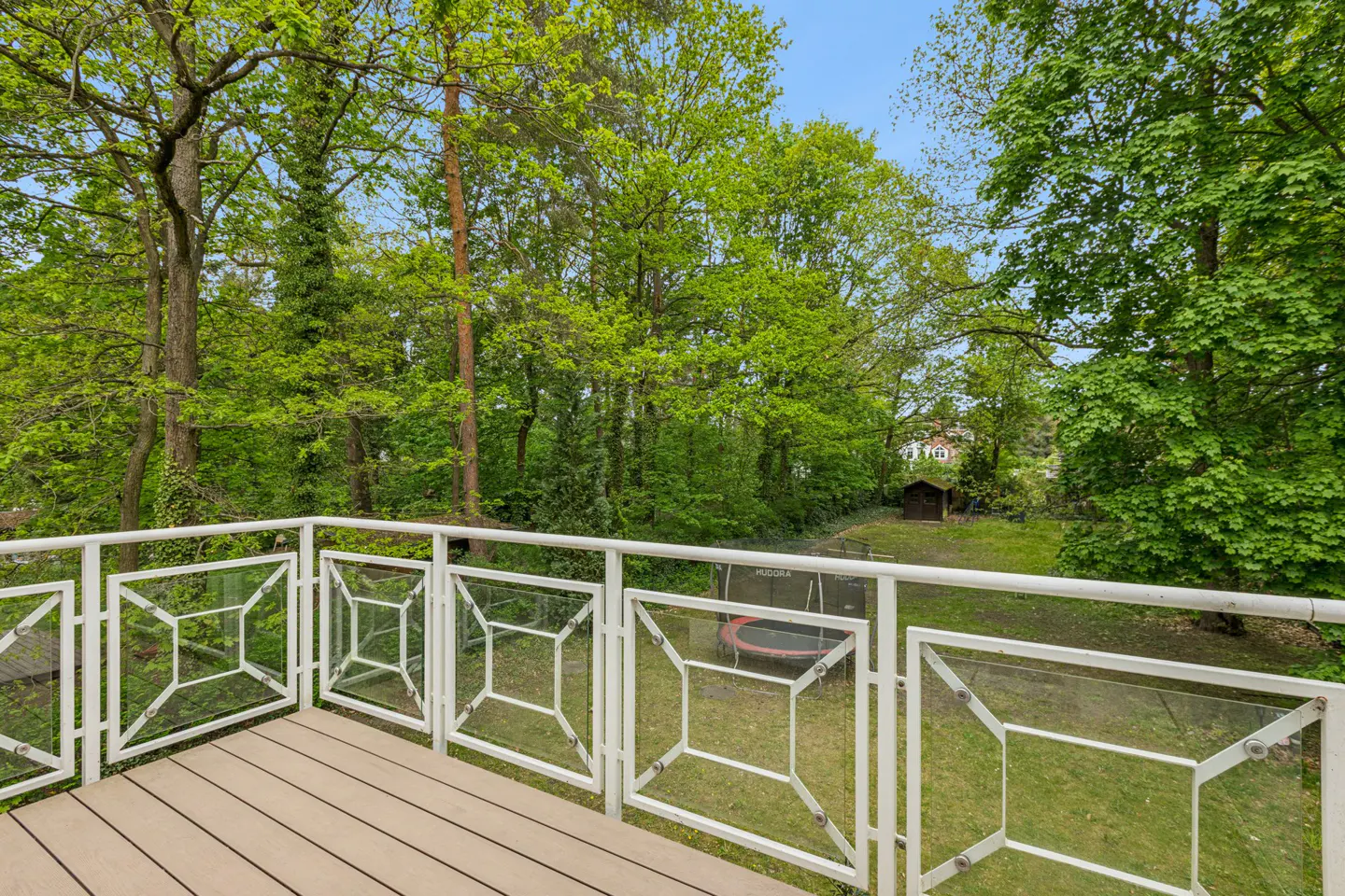 Balcony view with white railings, a wood floor, and a green lawn with trees. A trampoline and shed are in the background.