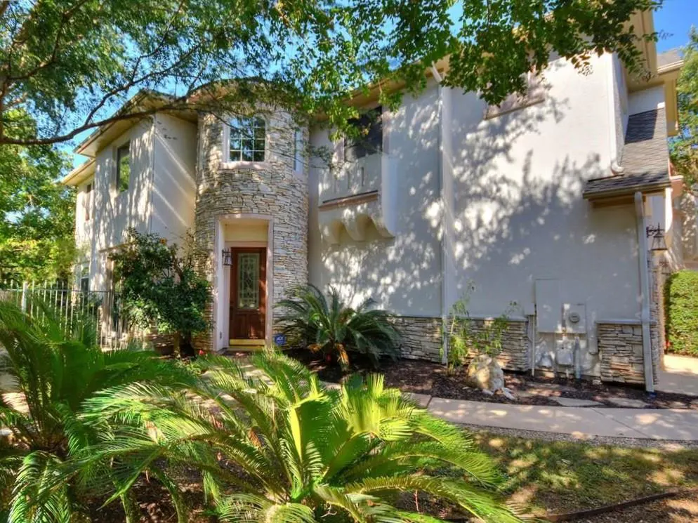 Two-story home with light stucco walls, stone accents, a dark wood front door, and lush green landscaping in the front.