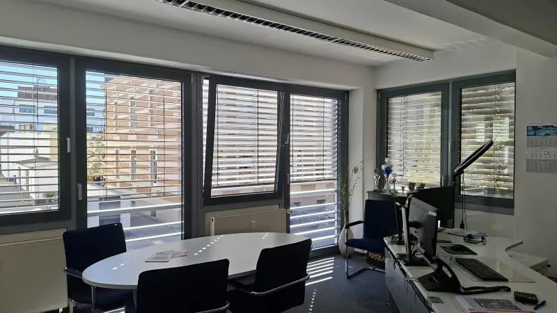 Bright office space with large windows, blinds, a white oval table with chairs, and a reception desk with computers.