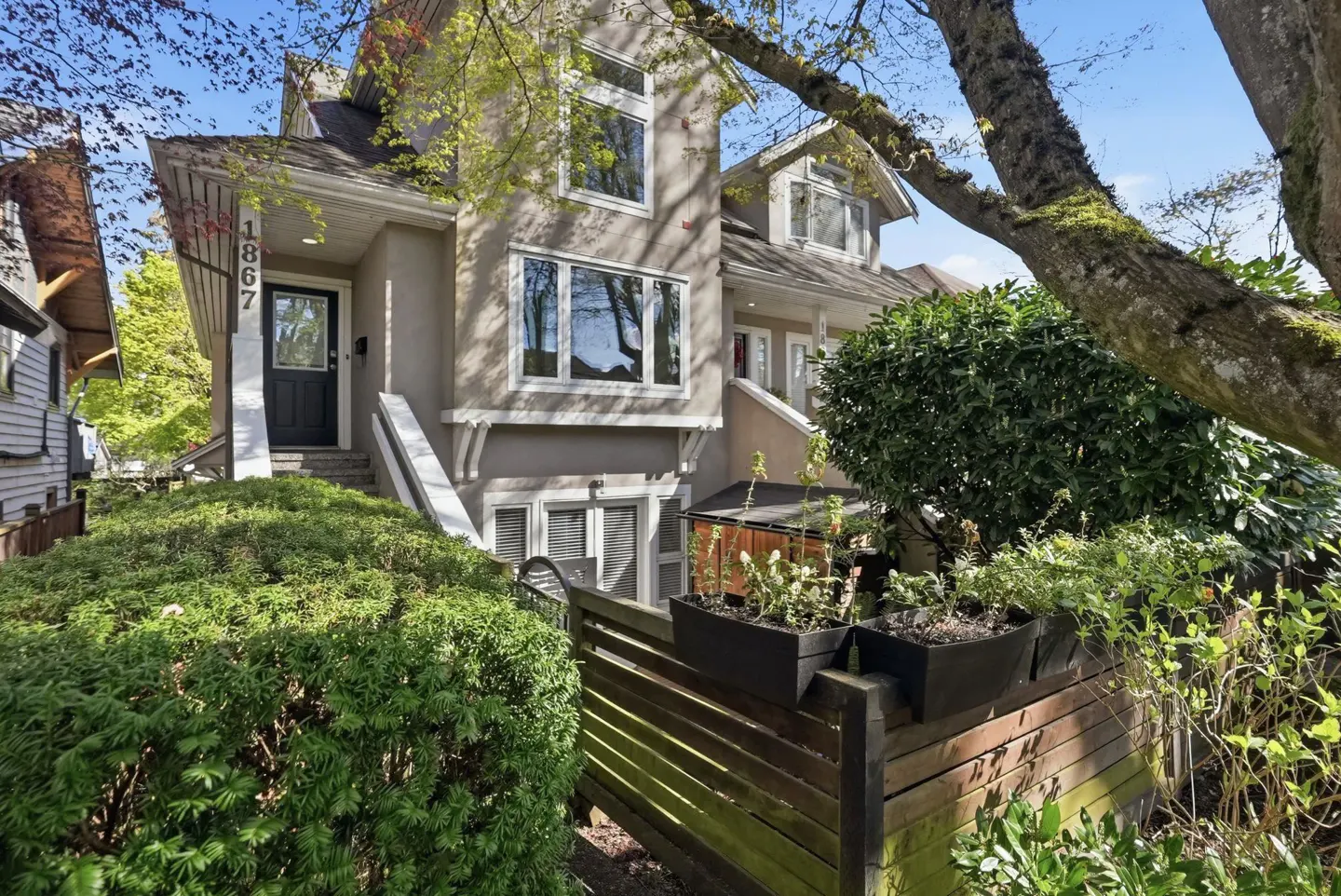 A two-story townhouse with a gray exterior, black door, and white trim, surrounded by greenery.