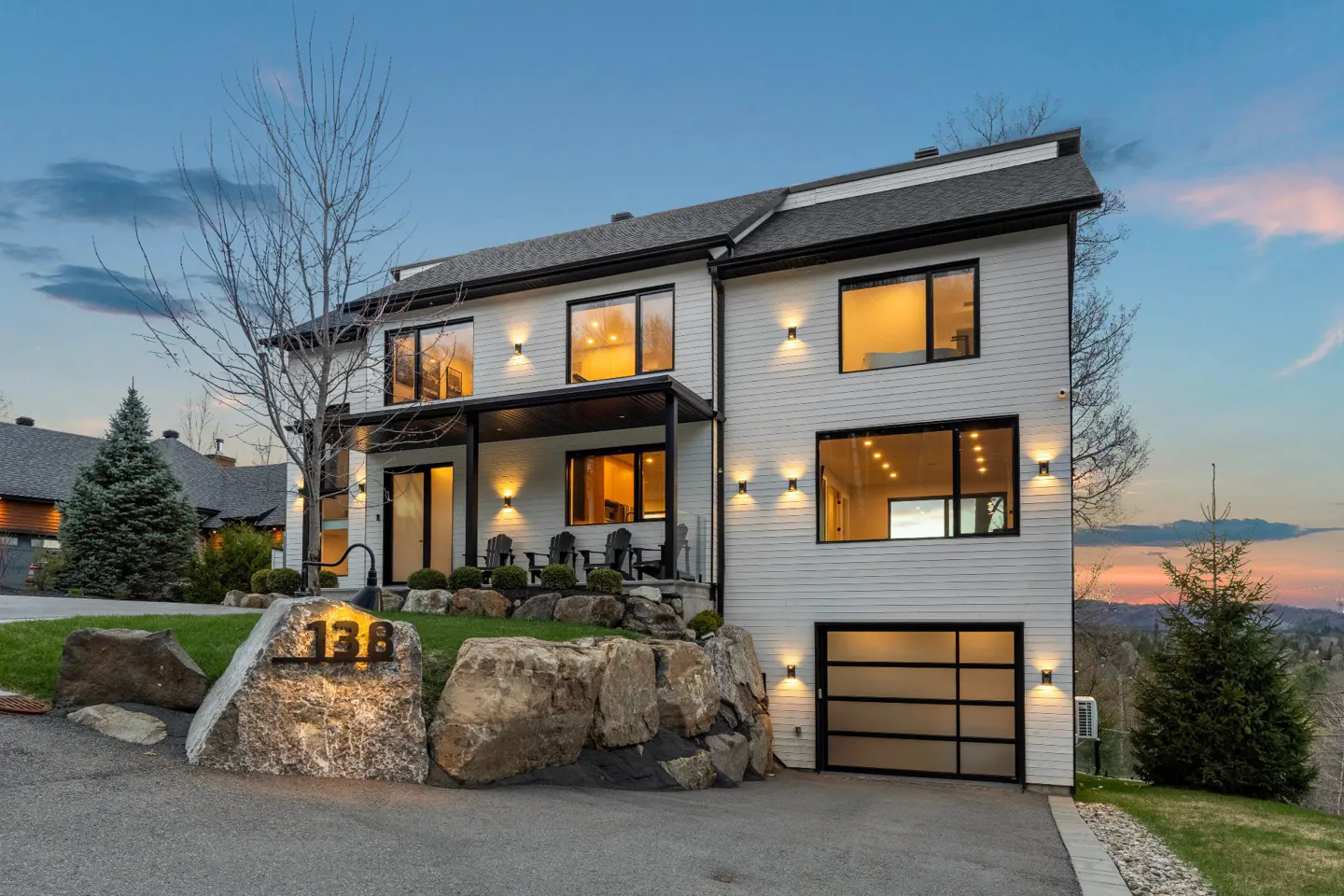 Modern two-story white house with black trim and a glass garage door at dusk. The house number, 138, is displayed on a large rock.