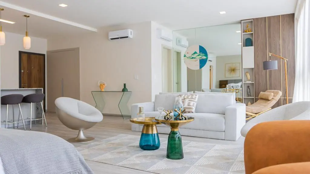 Bright, modern living room with white sofa, round chairs, and colorful glass tables on a geometric rug. A large mirror reflects the space.