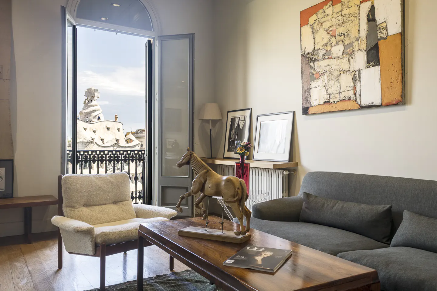 Living room with a gray sofa, a white armchair, and a wooden coffee table with a horse sculpture. Balcony doors open to a view of a building with a unique roof.