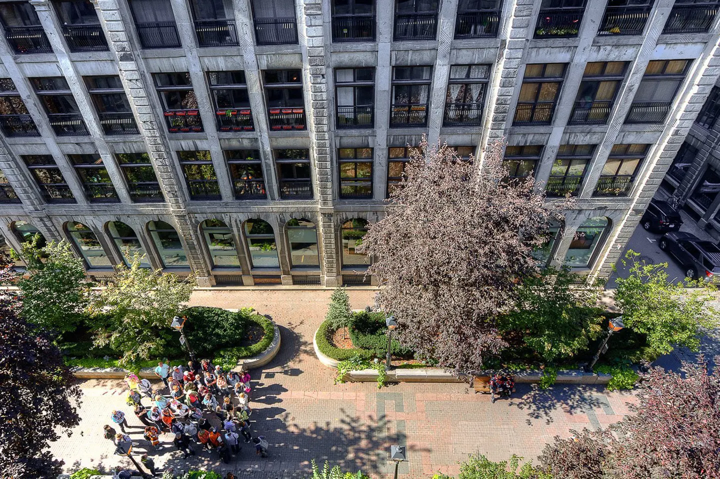 Aerial view of a gray stone building with arched windows, a brick courtyard, trees, and a group of people gathered below.