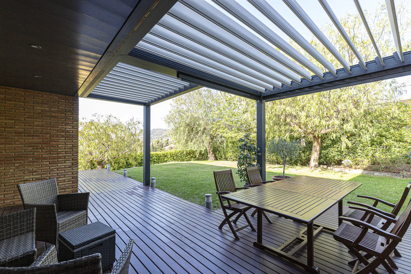 Outdoor patio with wooden deck, table, chairs, and pergola. Green lawn and trees in the background.