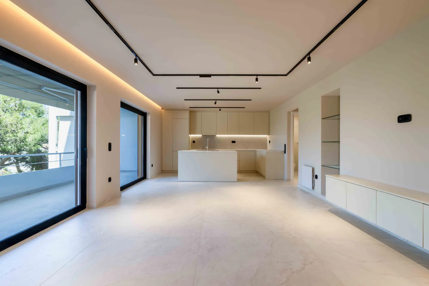 Bright, open-concept living space with a kitchen. Cream cabinets, island, and marble floors. Black track lighting and sliding glass doors to a balcony.