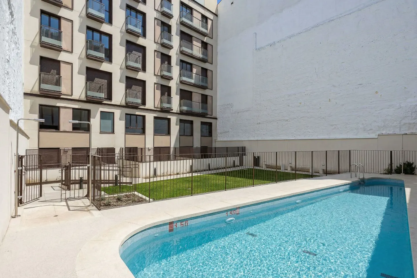 Apartment building with a pool. The pool is blue and rectangular. There is a small grassy area behind a black fence.