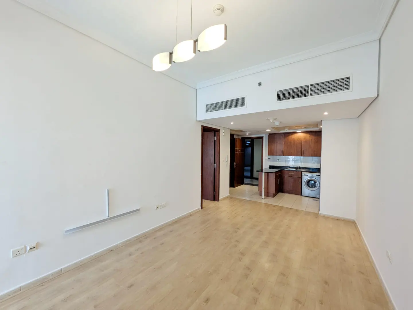 Empty living room with light wood floors, white walls, modern pendant lighting, and an open kitchen with brown cabinets.
