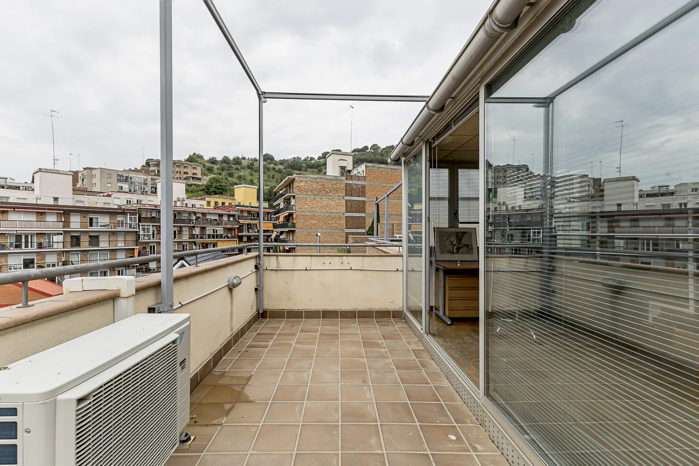 A tiled balcony with a view of buildings and a cloudy sky. An air conditioner sits on the left. Glass doors lead inside.