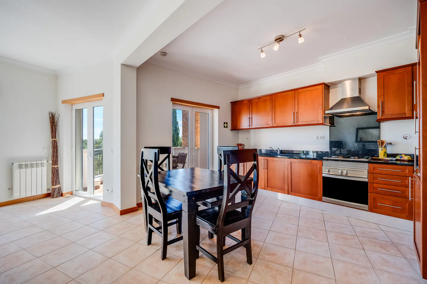 A bright kitchen with a dark wood table and chairs. The cabinets are wood with black countertops and stainless steel appliances. Tiled floor.
