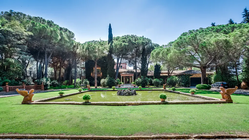 A grand villa with a central pond, stone fountain, and winged statues, surrounded by lush green lawns and tall pine trees.