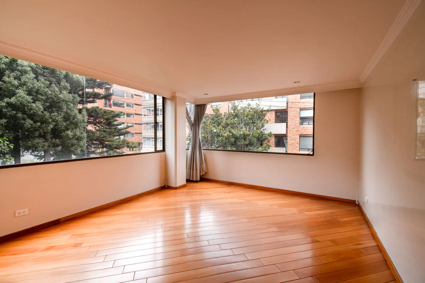 Empty room with light wood floors, white walls, and large windows. Outside, trees and brick buildings are visible.