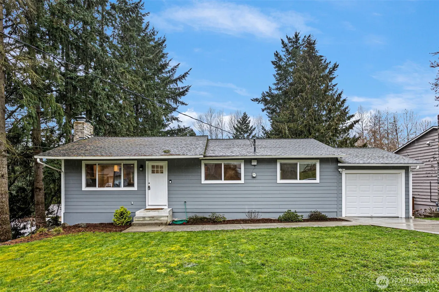 Single-story gray house with white trim, a white front door, and an attached garage, set against tall evergreen trees.