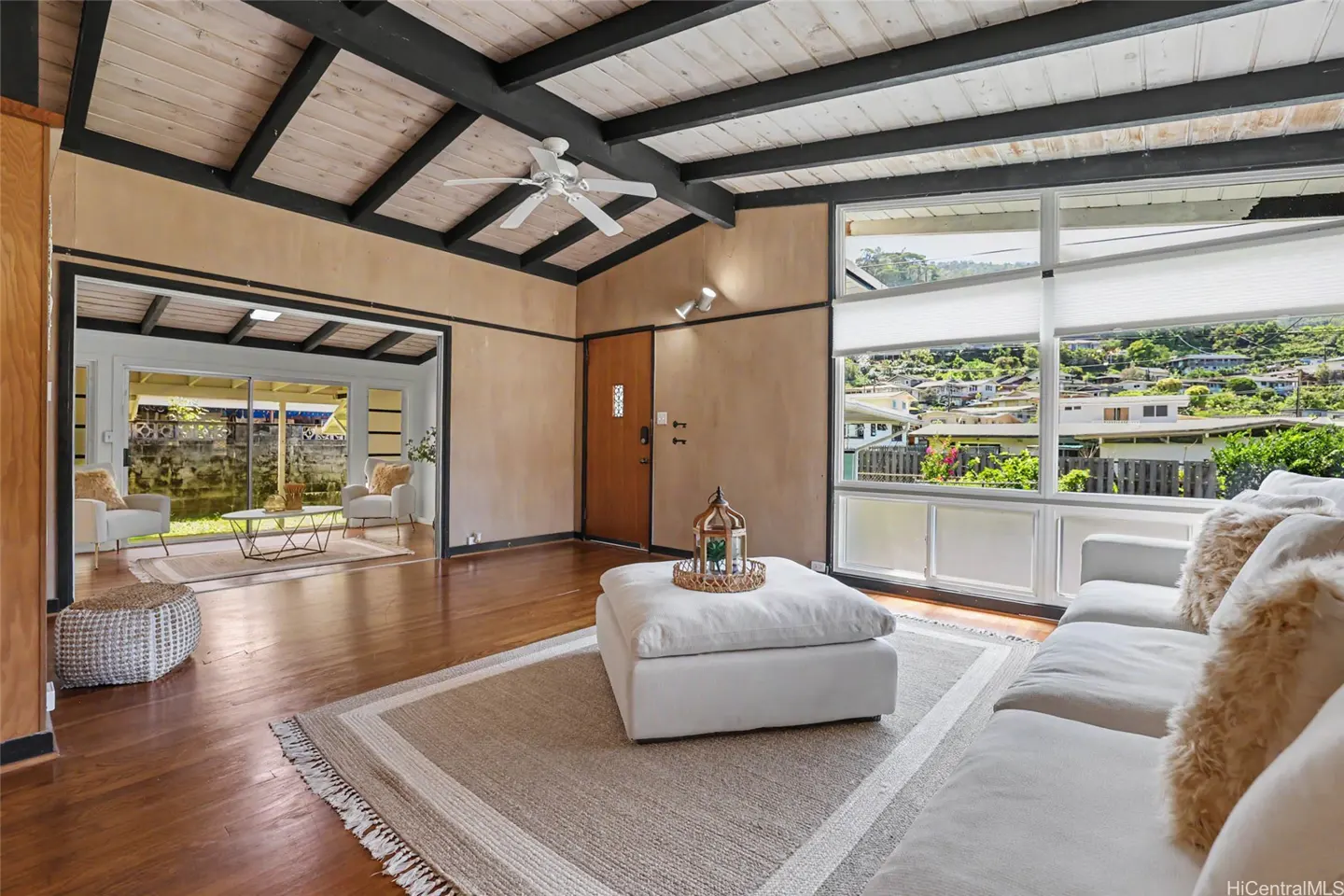 Living room with wood floors, white sofa, and ottoman on a rug. Large windows offer a view of a hillside neighborhood.