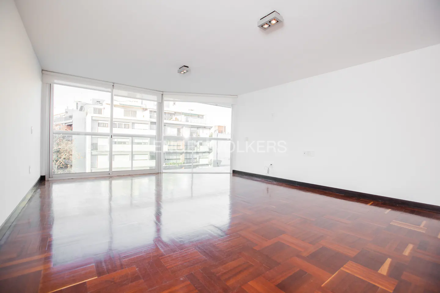 Empty room with herringbone wood floor, white walls, and large windows overlooking a city building.