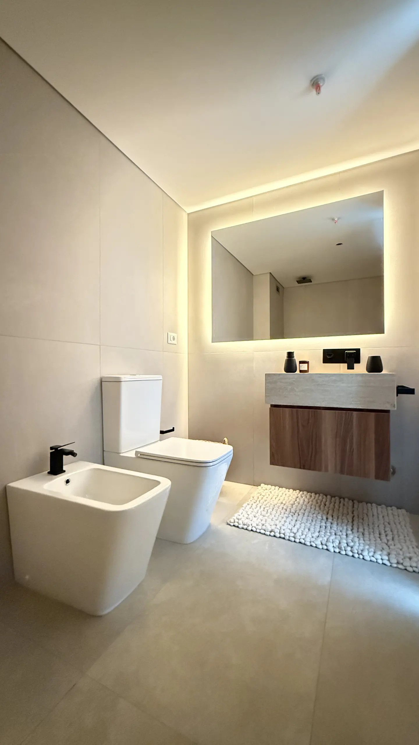 Bright bathroom with beige tile, white toilet and bidet with black fixtures. A wood vanity with a lighted mirror hangs above a white rug.