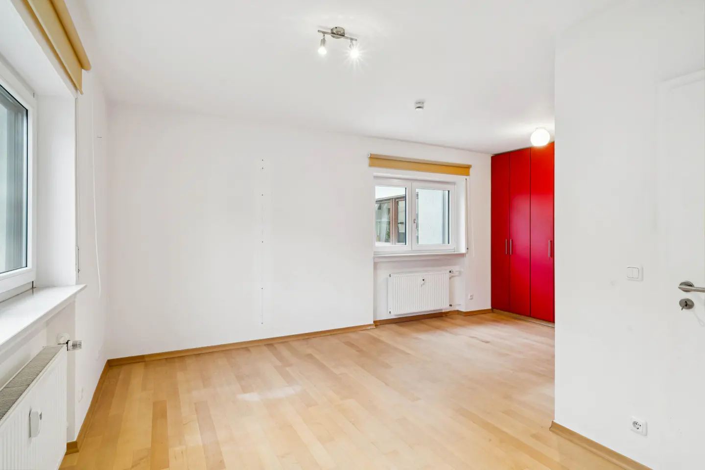 Bright, empty room with light wood floors, white walls, and two windows. A red wardrobe is on the right side of the room.