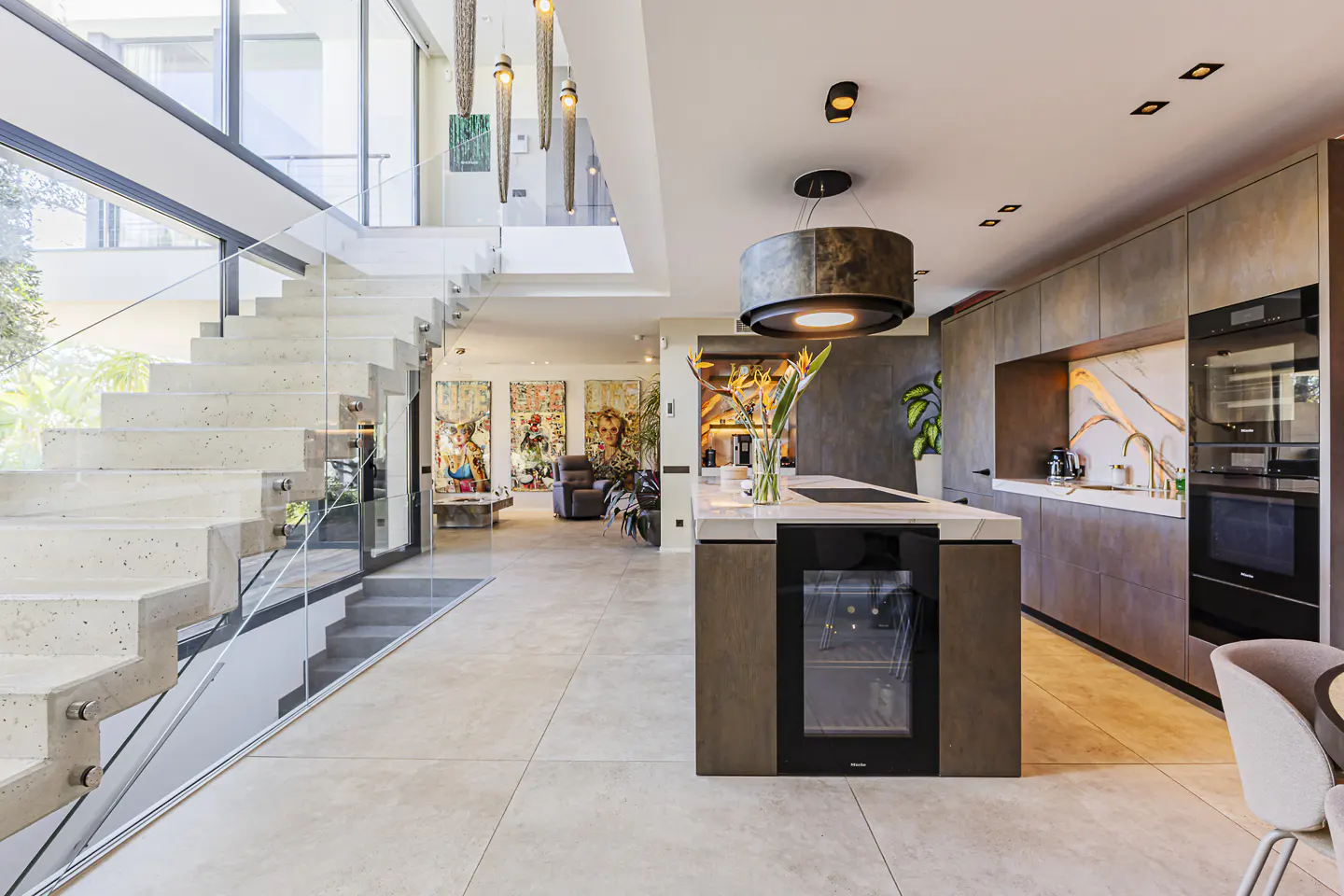 Open-concept home interior with a staircase, kitchen island, and living area. The kitchen has gray cabinets and a wine fridge. Art adorns the walls.