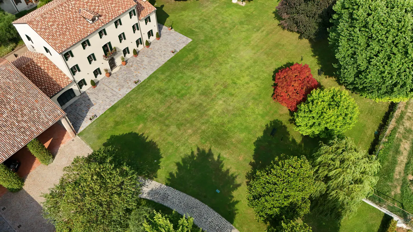Aerial view of a cream-colored house with a red tile roof, surrounded by a large green lawn and trees. A stone path curves through the yard.