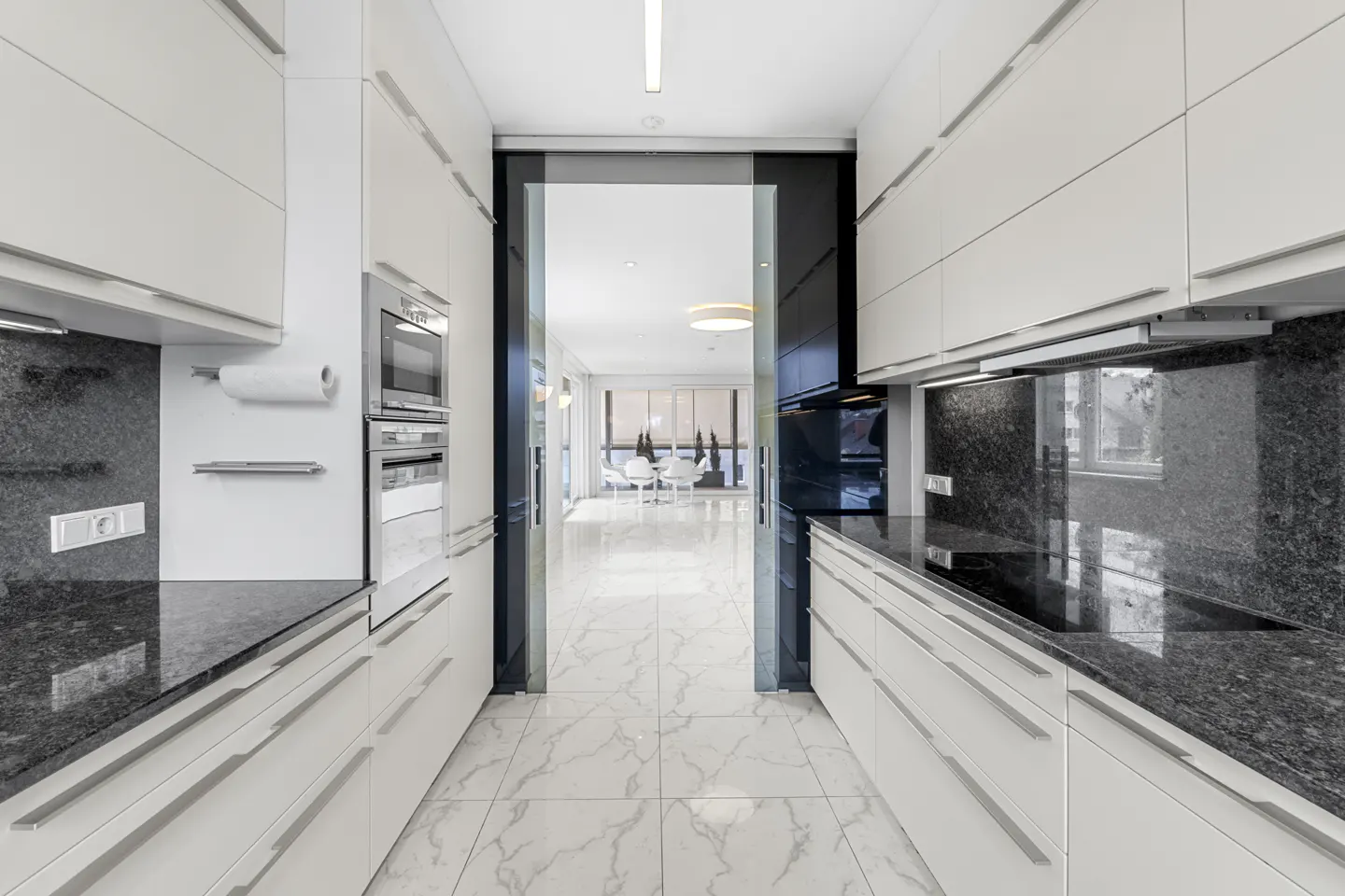 A modern kitchen with white cabinets, black granite countertops, and a marble floor. A doorway leads to a bright living area.