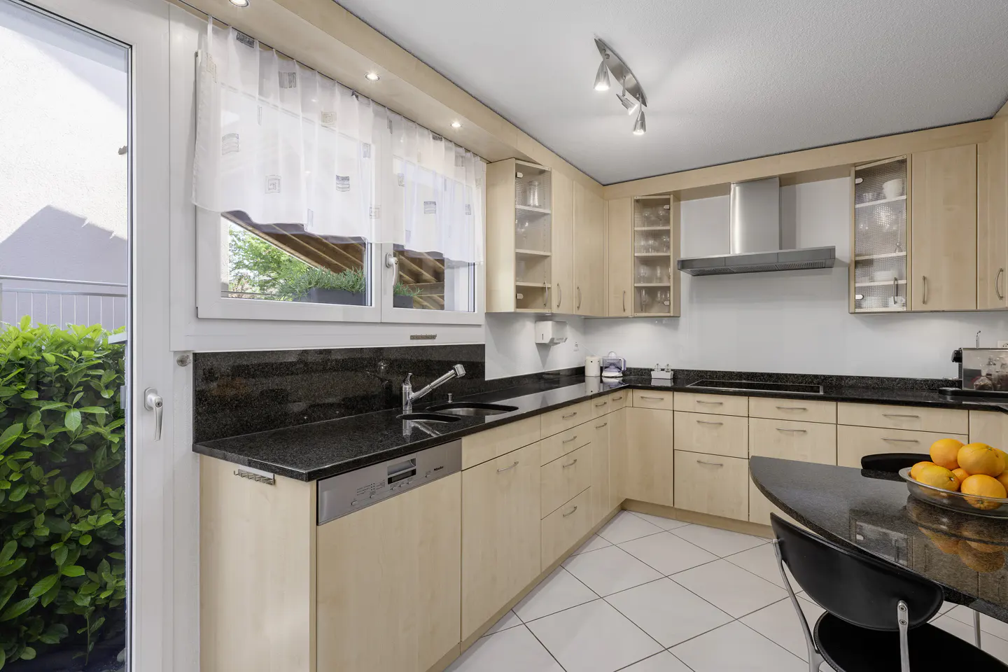 Bright kitchen with light wood cabinets, black granite countertops, and white tile floor. A black table with oranges sits in the corner.