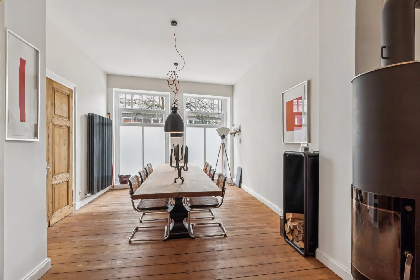 A dining room with a long wooden table, metal chairs, and a black pendant light. A wood-burning stove is in the corner.