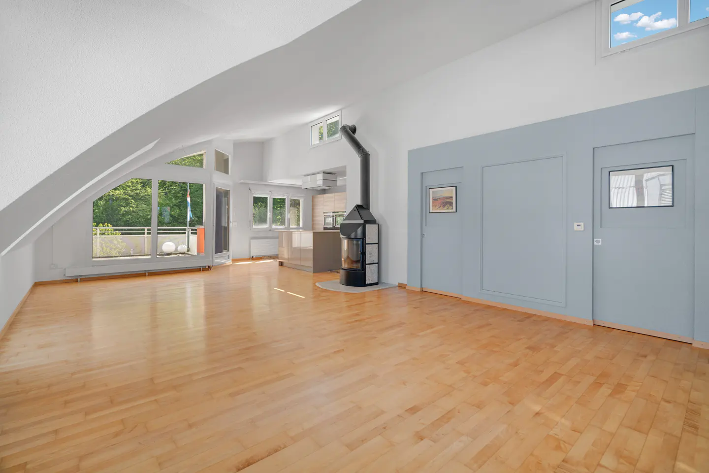 Bright, open living space with wood floors, a black fireplace, and a blue accent wall. Large windows offer a view of trees and a balcony.