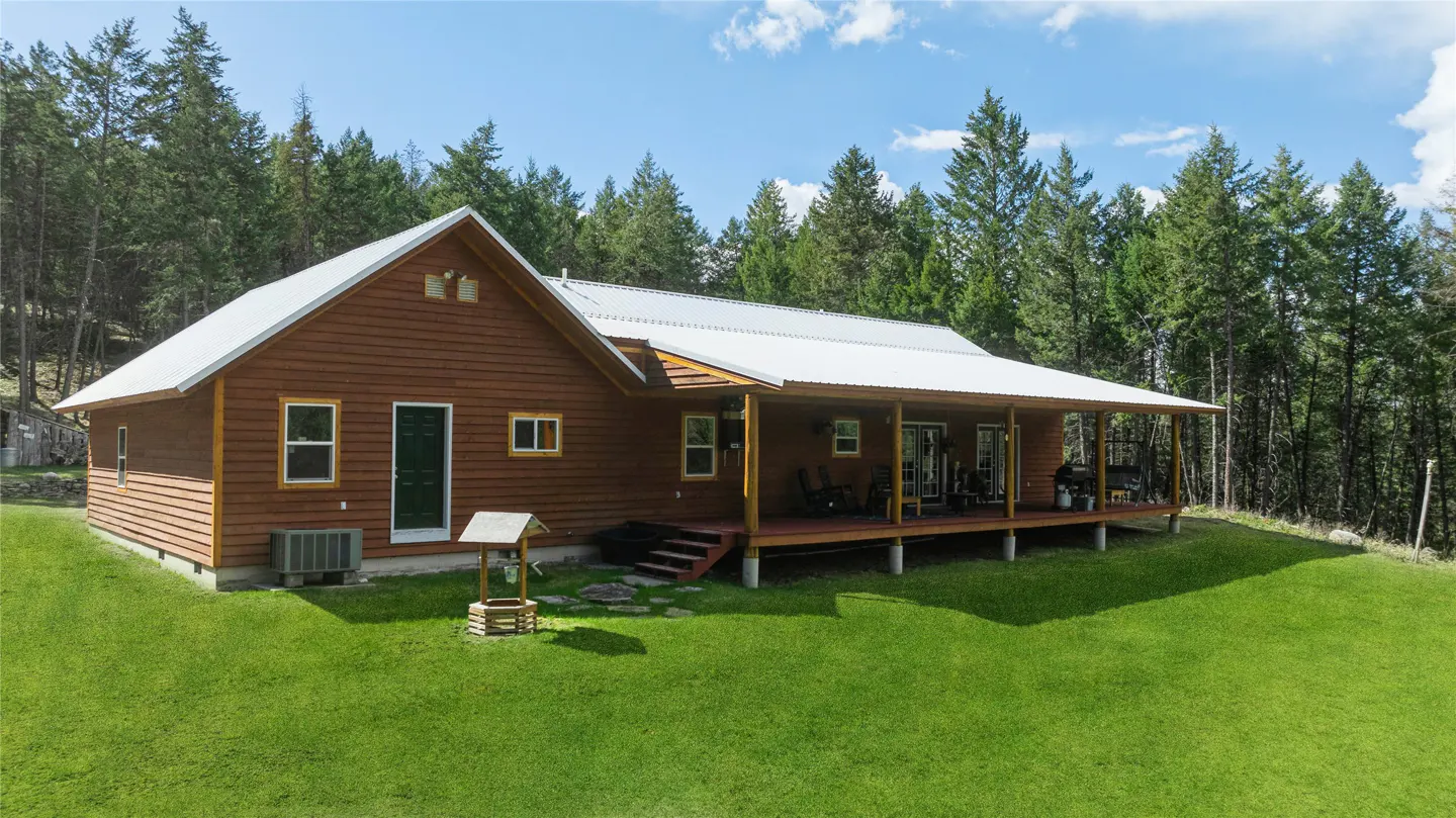 A brown, wood-sided house with a white roof and a long porch, set against a backdrop of green trees and a blue sky.