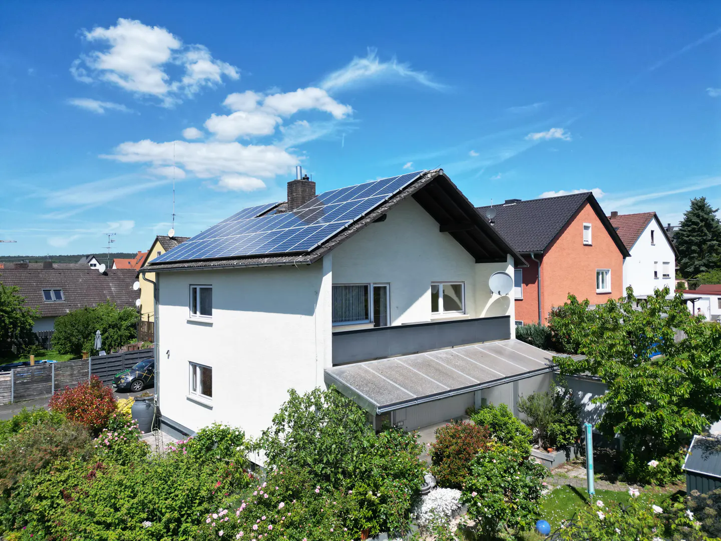 White house with solar panels on the roof, a balcony, and a lush garden under a clear blue sky in a residential area.