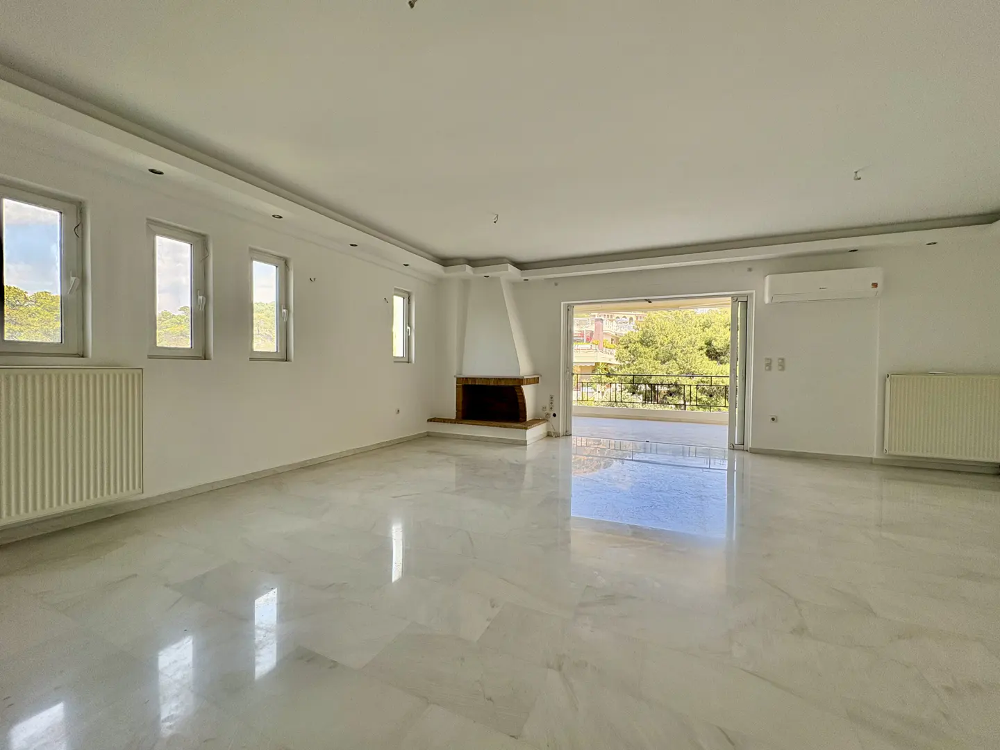 Bright, empty living room with marble floors, white walls, fireplace, windows, and balcony with green trees visible outside.