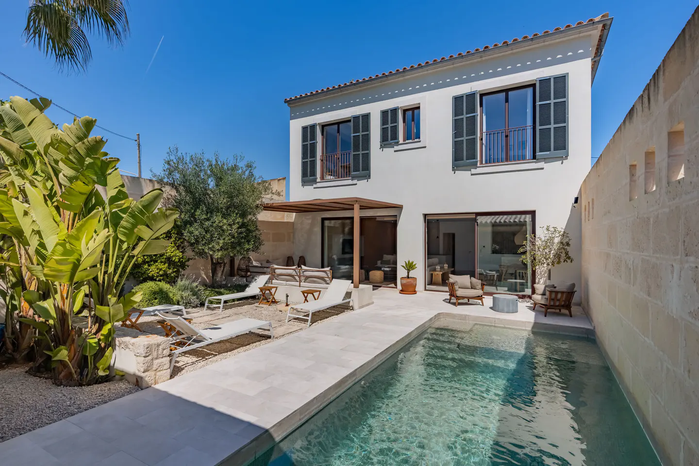 A white two-story house with a pool and patio area. Lounge chairs and tropical plants surround the pool. Blue sky.