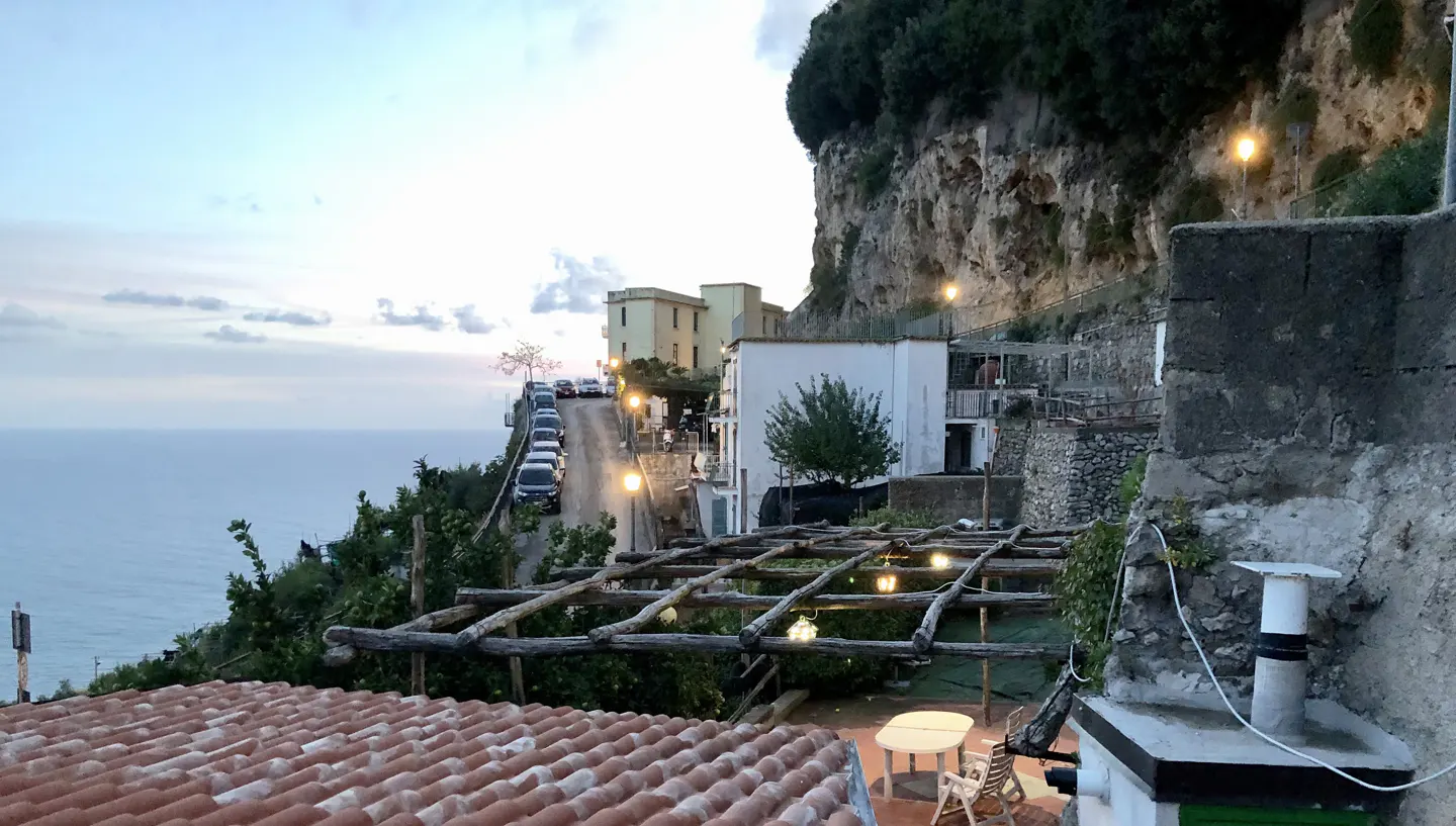 Coastal Italian town at dusk. Terracotta roof tiles in foreground, with a pergola, table and chairs. Ocean view, hillside homes, and cars parked on a steep road.
