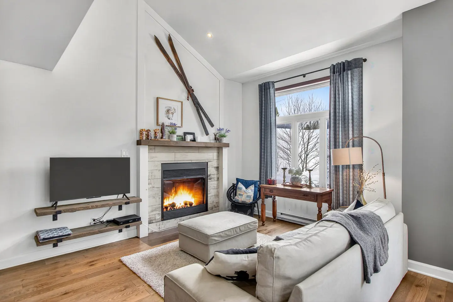 Cozy living room with a stone fireplace, burning fire, and skis mounted above. A white sofa and ottoman sit on a light rug. TV on wooden shelves.