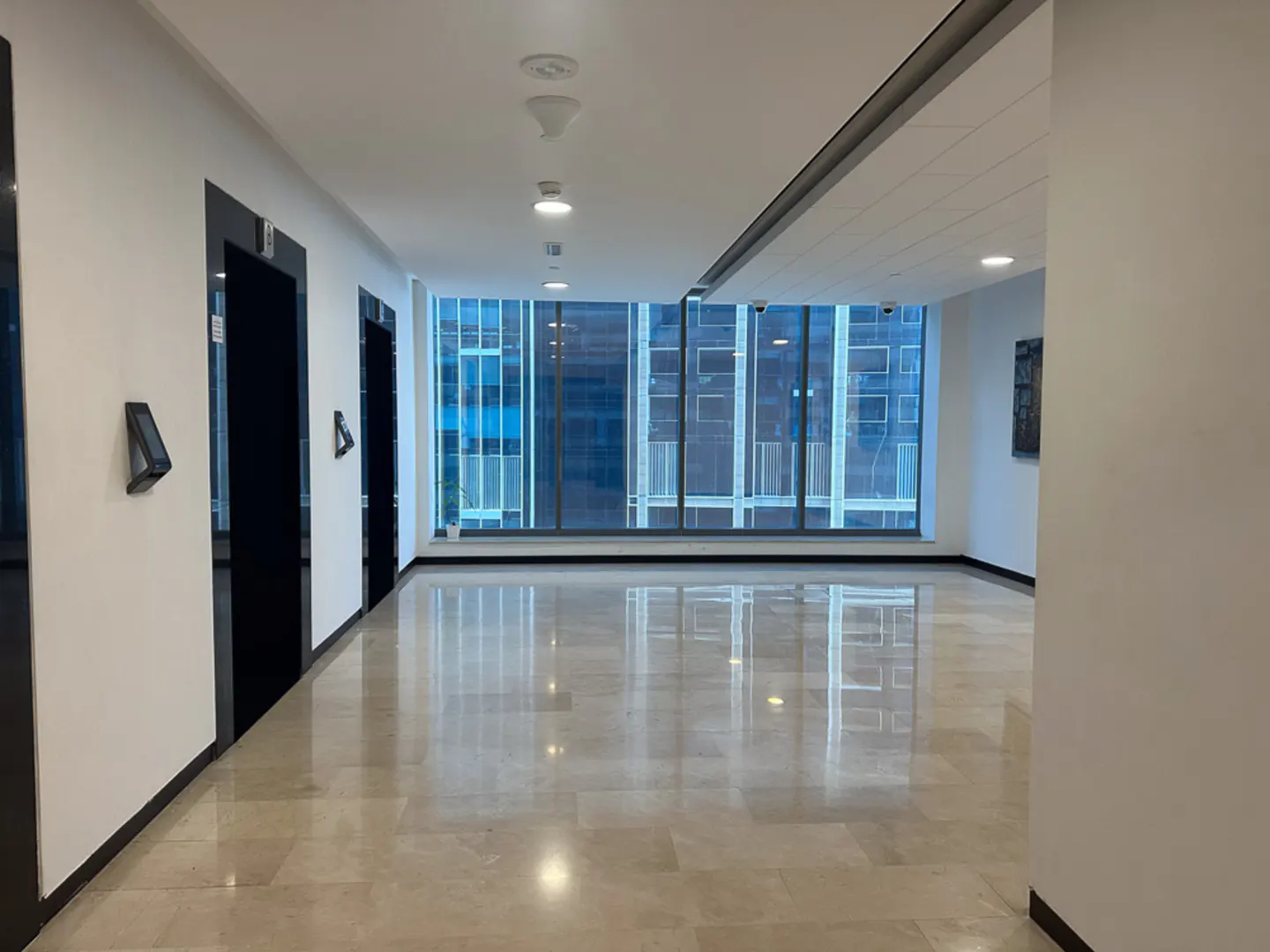 Hallway with marble floors, white walls, and black elevator doors. Large windows offer a city view.