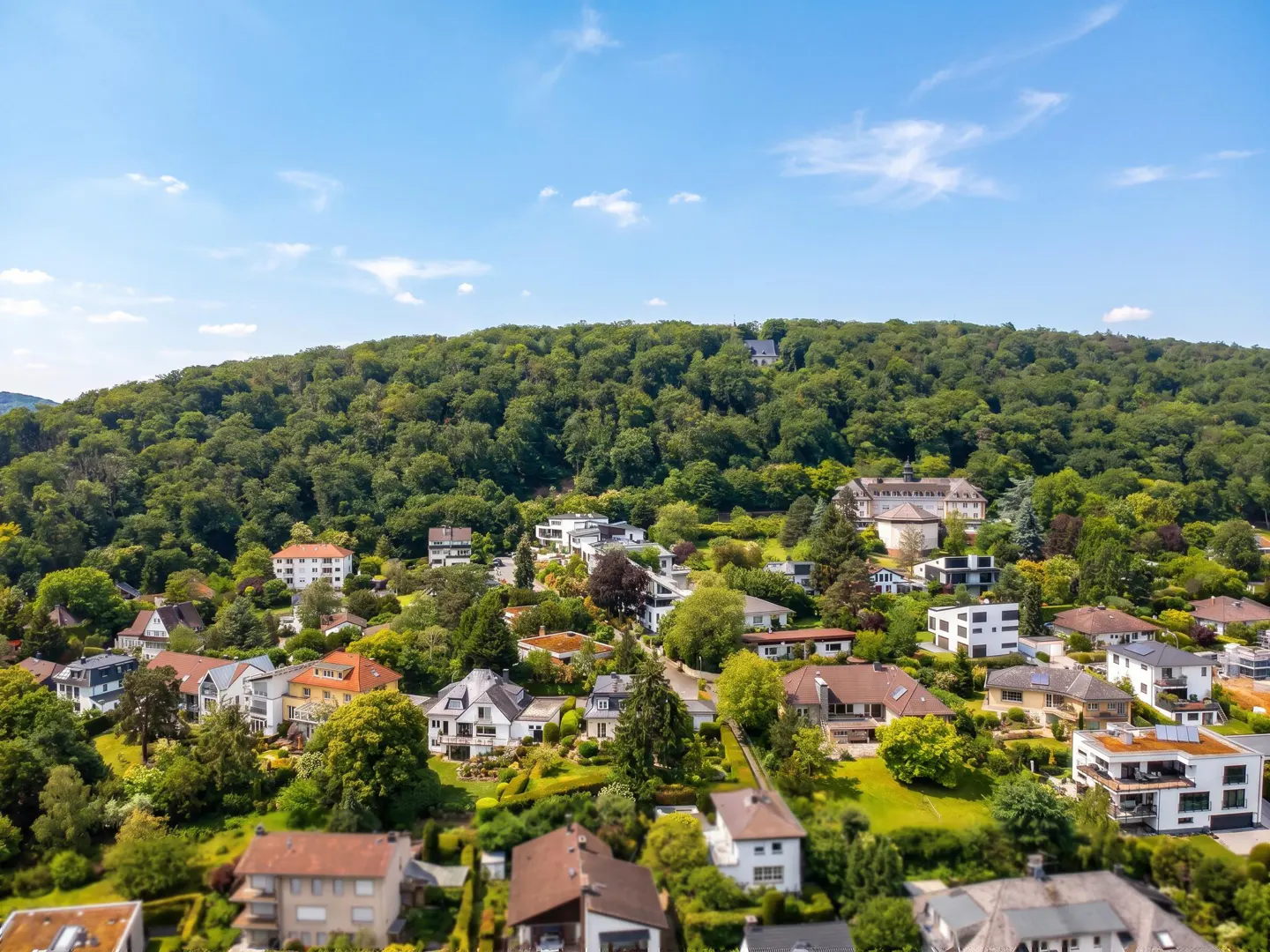 Aerial view of a residential neighborhood nestled at the base of a lush, green forested hill under a bright blue sky.