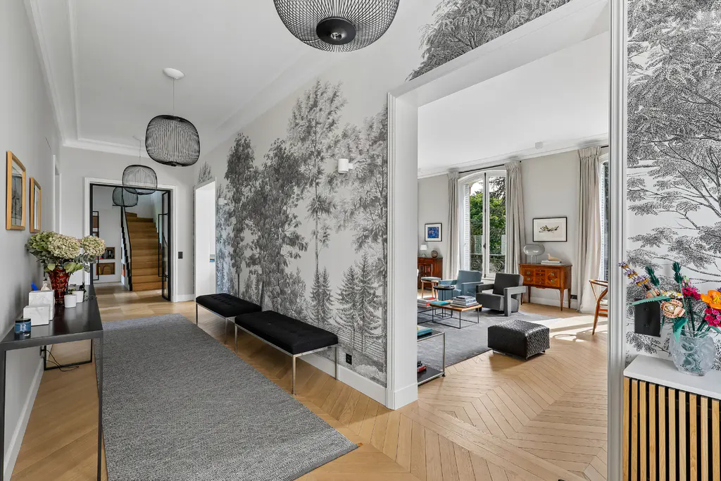 A bright hallway with wood floors, a gray rug, and a black console table with flowers. A black and white forest mural adorns the wall. A living room is visible through an archway.