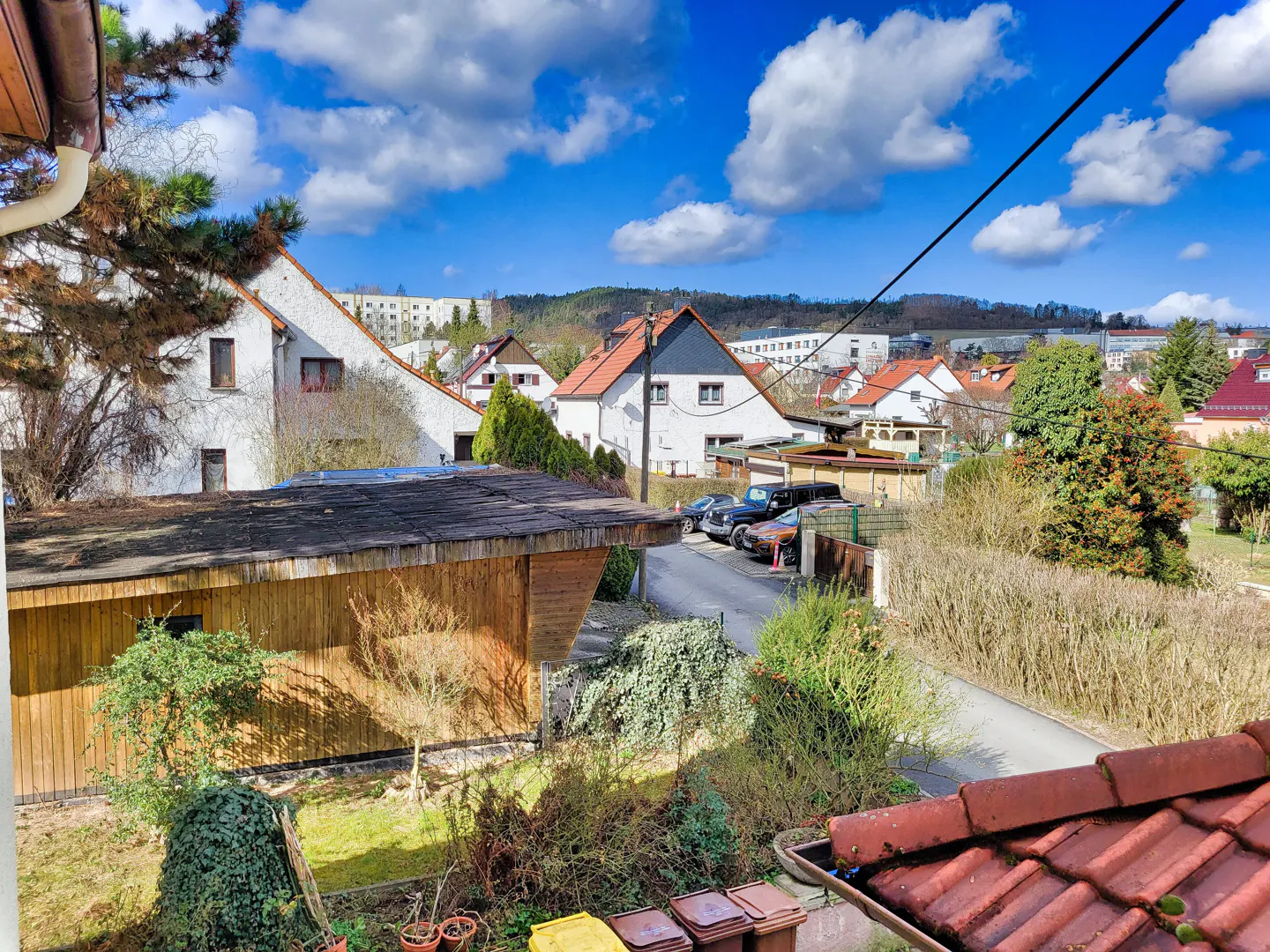 View of a European neighborhood with white houses, red roofs, and a blue sky with fluffy clouds. A wooden shed and garden are in the foreground.
