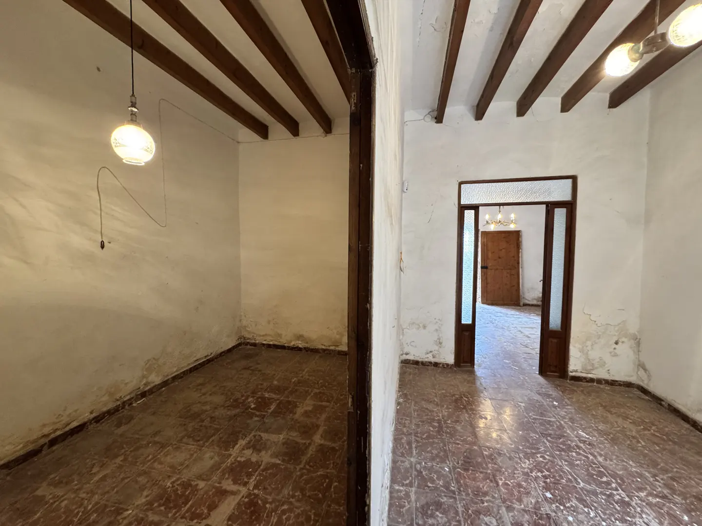 Interior view of two rooms with white walls, brown ceiling beams, and red tile floors, separated by a dark wood frame.
