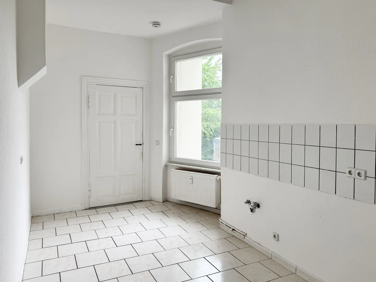 Bright, empty kitchen with white walls, tiled floor, and a window. A white door is on the left, and a tiled backsplash is on the right.