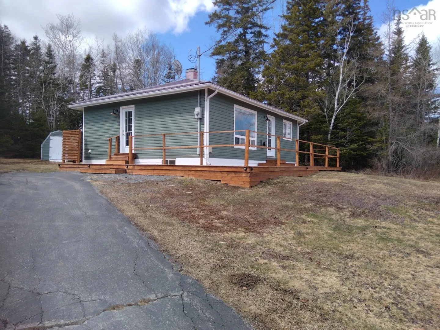 A one-story green house with a wooden deck and railing, set against a backdrop of trees and a blue sky.