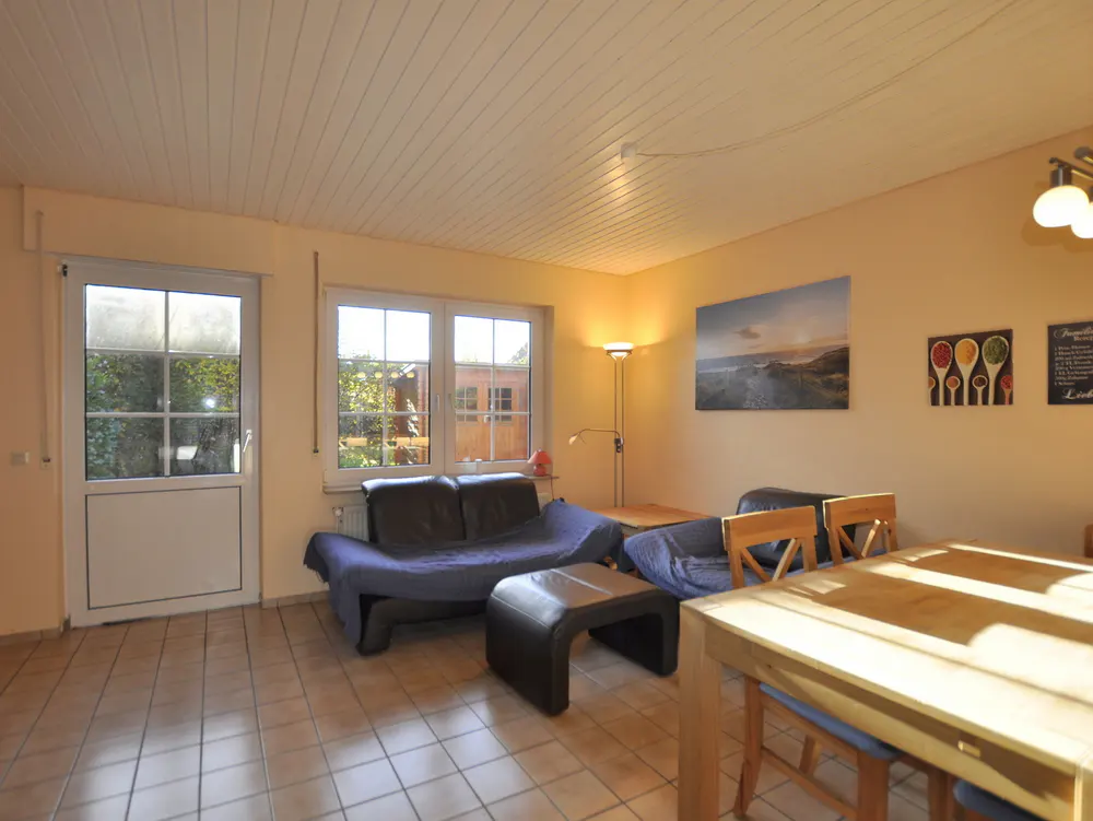 Living room with tiled floor, two dark sofas with blue throws, a wooden dining table, and a glass door leading outside.