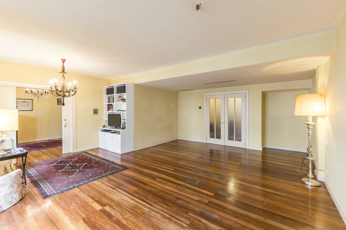 A spacious living room with hardwood floors, yellow walls, and a chandelier. A white bookcase and double doors are visible.