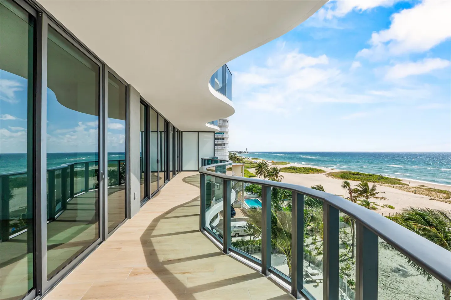 A curved balcony with glass railings overlooks a sandy beach and blue ocean under a partly cloudy sky.