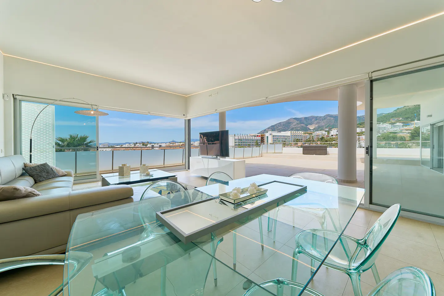 Bright, modern living room with glass table and chairs. Balcony with ocean view and mountains in the distance. Cream sofa and white walls.