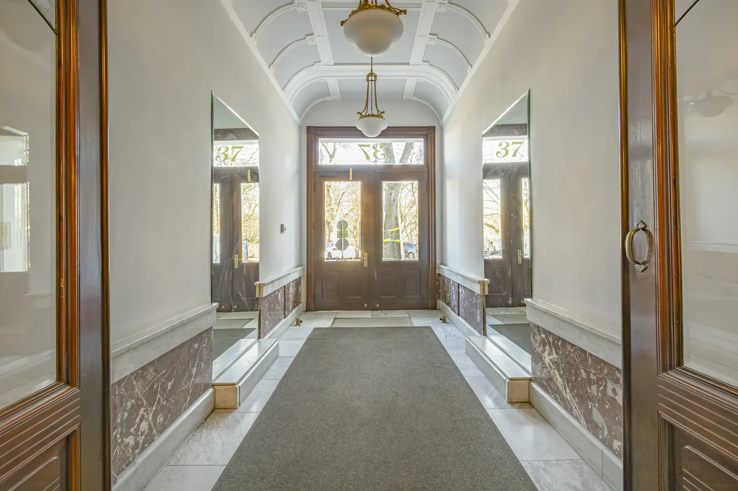 Hallway with marble walls, a gray carpet, and a dark wood door at the end. Mirrors on the walls reflect the door. Two hanging lights illuminate the space.